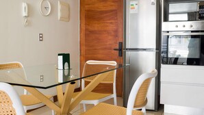 A modern kitchen with a glass-top table, white chairs, a refrigerator, and a wooden door. - Modern Chic Oasis: Sophisticated Las Condes Stay (Las Condes)