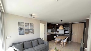 A modern living room with a grey sofa, a dining table with chairs, and a kitchen area with dark cabinets and a wooden door. - Viña del Mar: Apartment with sea views and balcony (Viña del Mar)