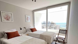 A modern bedroom with two beds, a wooden chair, and a large window offering a city view. - Viña del Mar: Apartment with sea views and balcony (Viña del Mar)