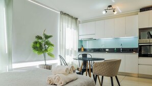 Una habitación de hotel moderna con cama, una planta en maceta, una mesa de comedor con sillas y una kitchenette.