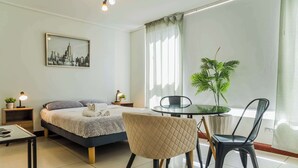 A modern bedroom with a bed, bedside tables, a round table, chairs, a potted plant, and a cityscape painting on the wall. - Charming Studio in Las Condes with Pool | Ideal 2 (Las Condes)