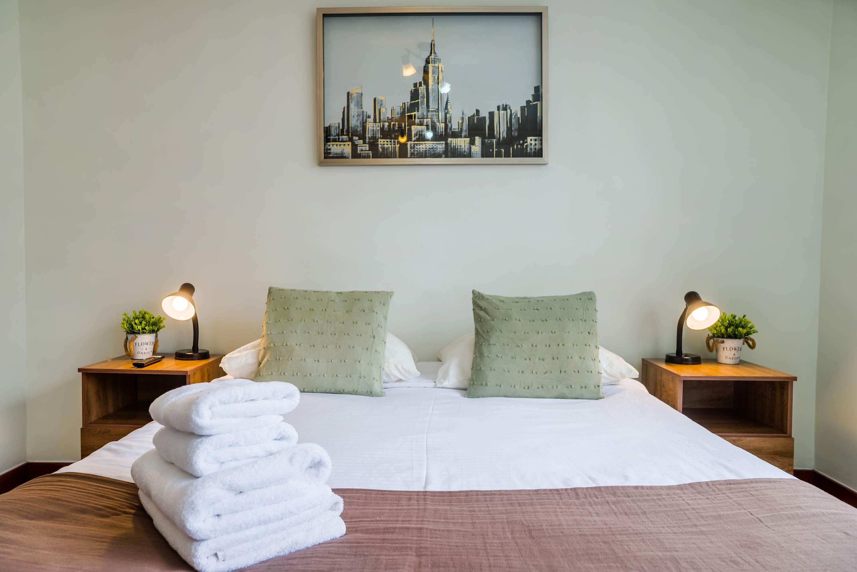 A hotel room with a bed, two nightstands, a framed cityscape picture, and a stack of white towels.