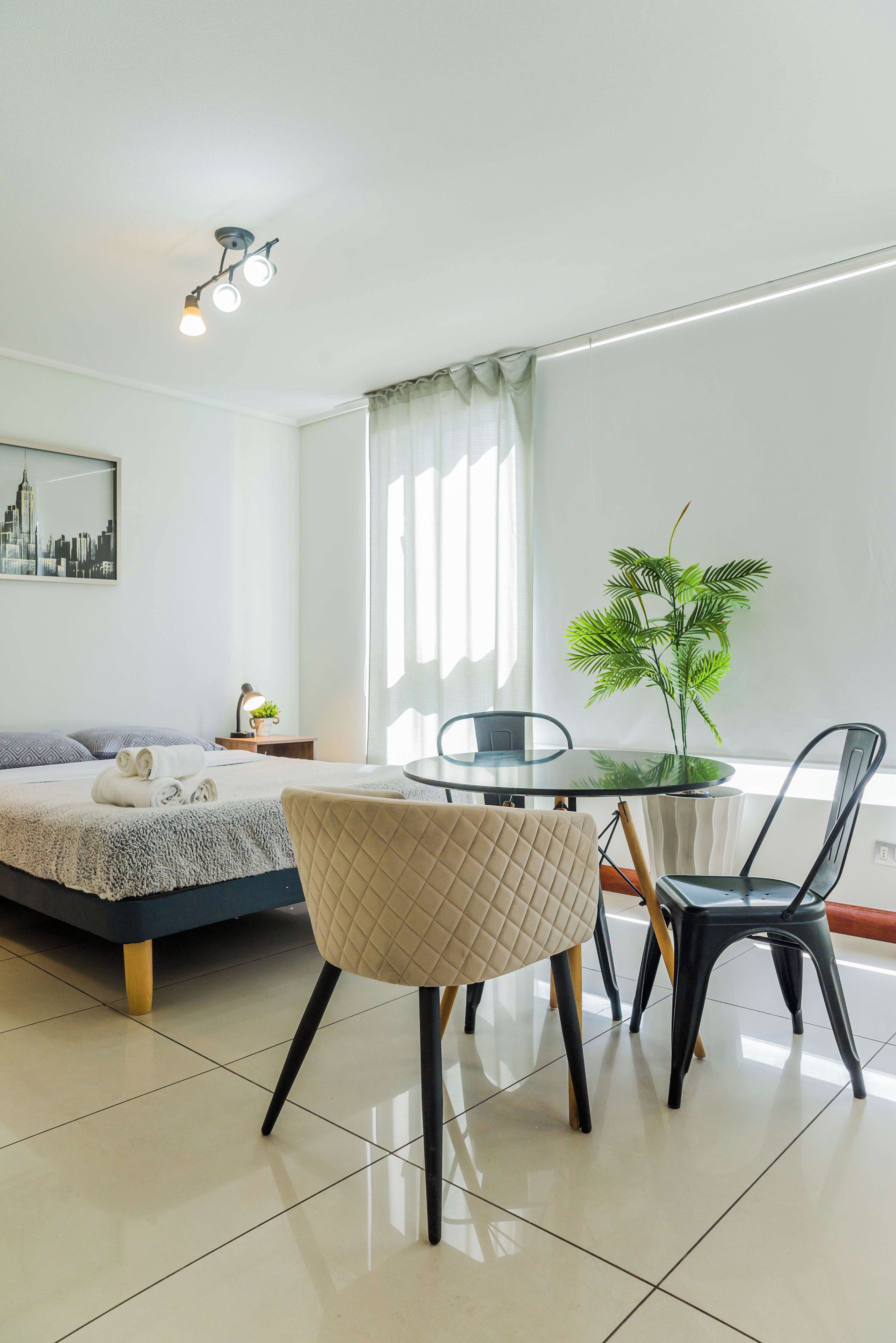 A modern hotel room with a bed, a round glass table, two chairs, a potted plant, a lamp, and a cityscape painting on the wall.