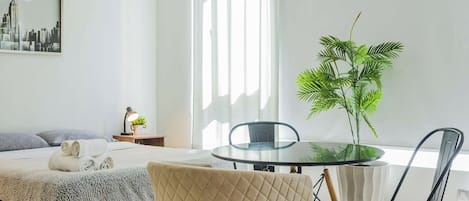 A modern hotel room with a bed, a round glass table, two chairs, a potted plant, a lamp, and a cityscape painting on the wall.