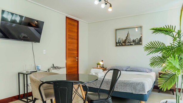 Una habitación de hotel con una cama, un televisor fijado a la pared, una mesa de comedor con sillas, una planta en maceta y un cuadro de un paisaje urbano.
