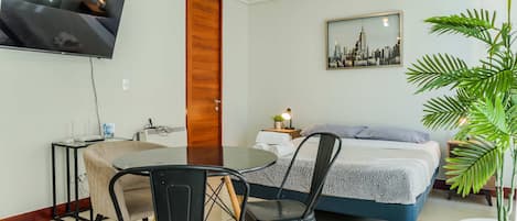 A hotel room with a bed, a TV mounted on the wall, a dining table with chairs, a potted plant, and a cityscape painting on the wall.