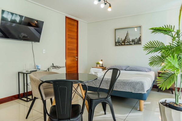 Une chambre d’hôtel avec un lit, un téléviseur fixé au mur, une table à manger avec des chaises, une plante en pot et un tableau représentant un paysage urbain accroché au mur.