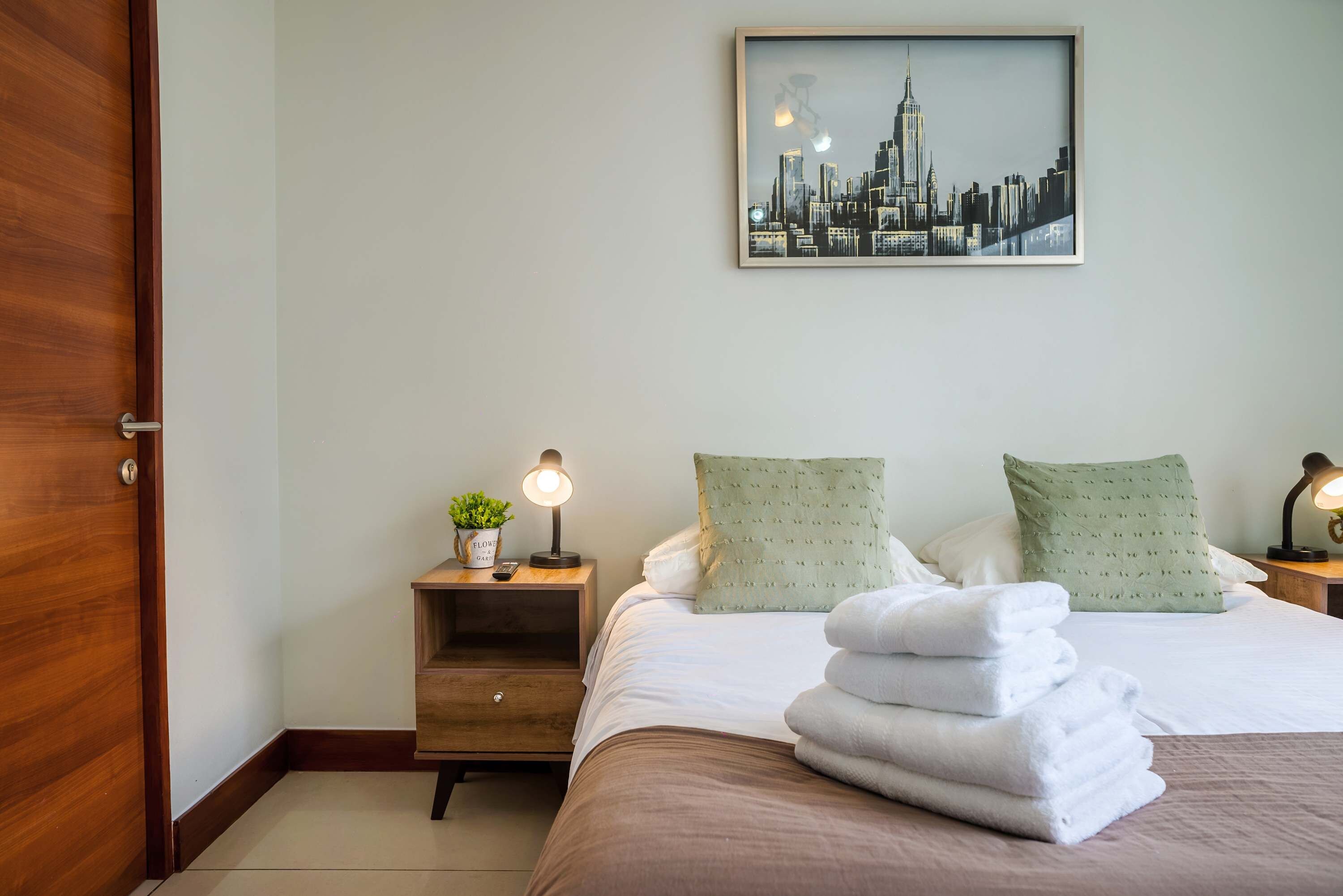A hotel room with a bed, two bedside tables, a nightstand, a framed cityscape picture, and a door.
