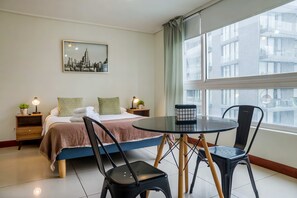 A hotel room with a bed, two bedside tables with lamps, a small table with chairs, a window with curtains, and a cityscape painting on the wall.
