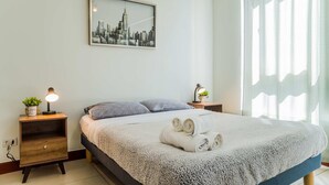 Una cama bien tendida con toallas enrolladas, un cuadro de la ciudad en la pared y una mesita de noche con una lámpara y una planta pequeña.