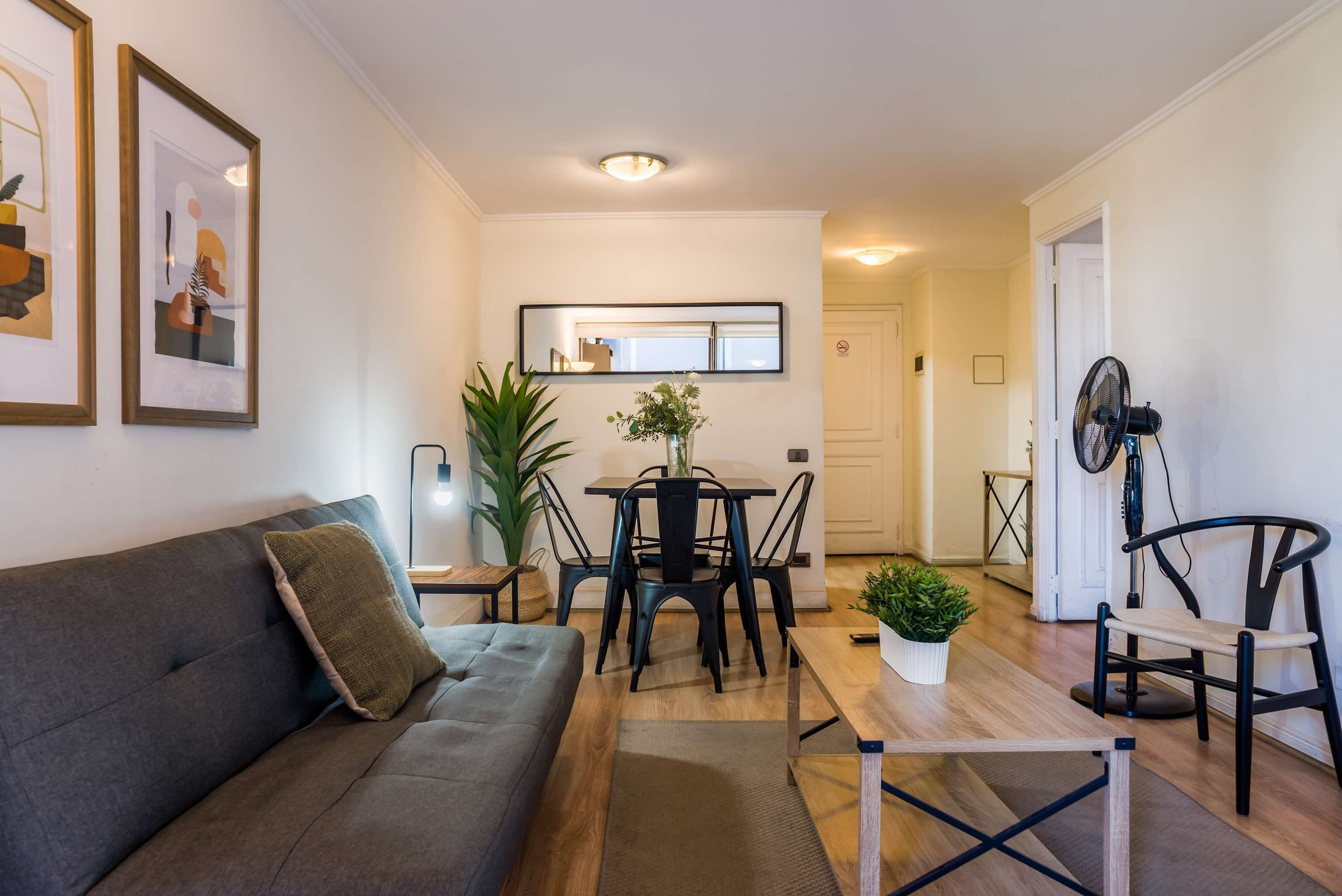 A living room with a sofa, a dining table with chairs, a small table with a plant, and a fan.