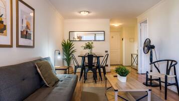 A living room with a sofa, a dining table with chairs, a small table with a plant, and a fan.