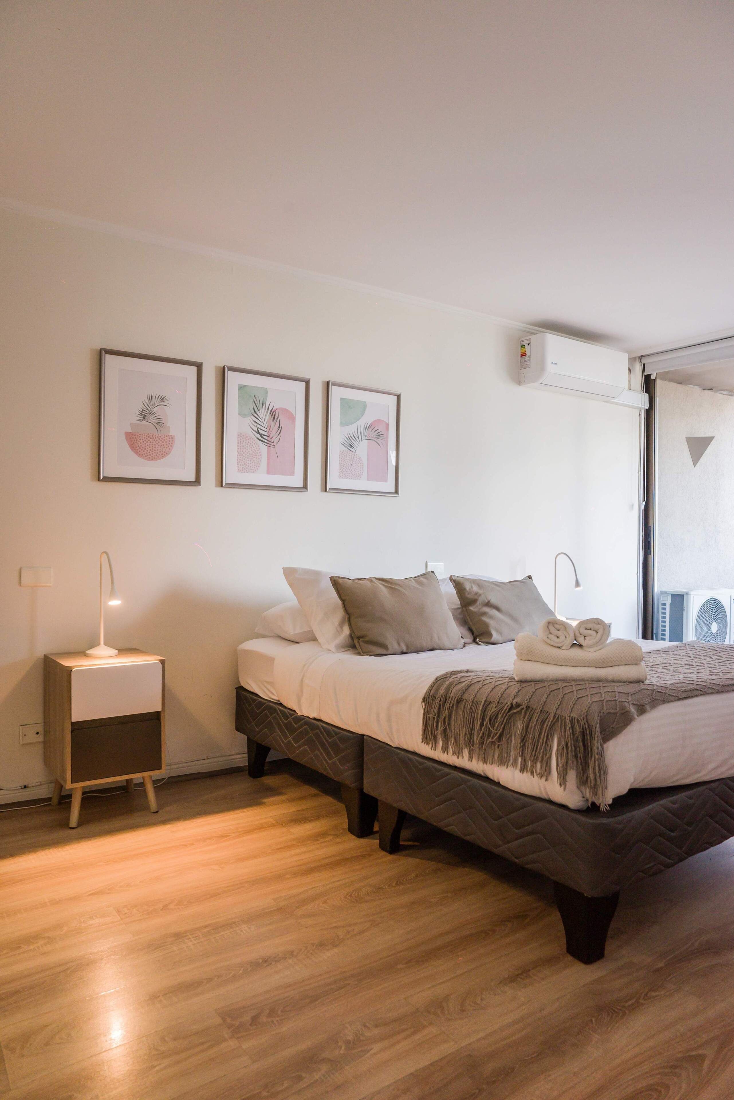 A bedroom with a bed, bedside tables, a wall-mounted air conditioner, and framed artwork on the wall.