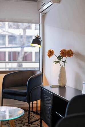 A room with a desk, a chair, a glass table, a vase with flowers, and a window with blinds.