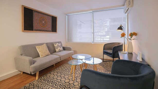 A living room with a sofa, two round glass tables, a chair, a vase with flowers, and a large window. - Comfortable 1BR/1BA in Las Condes | Prime Location (Las Condes)