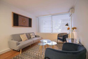 Ein Wohnzimmer mit einer Couch, zwei runden Glastischen, einem Sessel, einer Vase mit Blumen und einem großen Fenster.