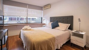 A bedroom with a bed, bedside table, lamp, and a window with blinds. - Comfortable 1BR/1BA in Las Condes | Prime Location (Las Condes)