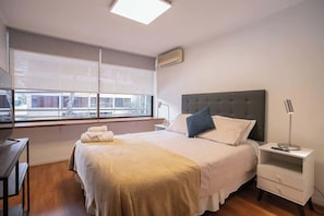 A bedroom with a bed, bedside table, lamp, and a window with blinds.