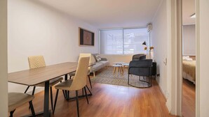 A modern living room with a wooden dining table, two sofas, a coffee table, and a view of the city through large windows. - Comfortable 1BR/1BA in Las Condes | Prime Location (Las Condes)