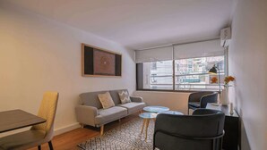 A modern living room with a sofa, a dining table, a coffee table, and a window with a view of buildings. - Comfortable 1BR/1BA in Las Condes | Prime Location (Las Condes)