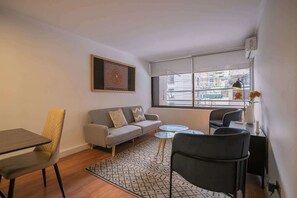A modern living room with a sofa, a dining table, a coffee table, and a window with a view of buildings.