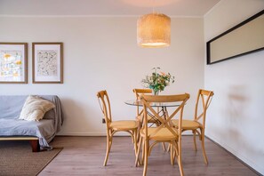 A dining area with a round table, four wooden chairs, a hanging light fixture, and two framed artworks on the wall.