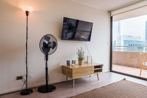 A modern living room with a flat-screen TV mounted on the wall, a standing fan, a wooden console table with decorative items, and a view of the city through a large window.
