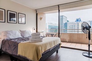 Una habitación de hotel moderna con una cama grande, un balcón con vistas a la ciudad y un ventilador de pie.
