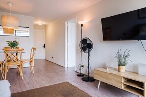 A modern living room with a flat-screen TV, a standing fan, a dining area with a table and chairs, and a potted plant. - Chic 1BR Oasis in Las Condes: Pool and City Views (Las Condes)