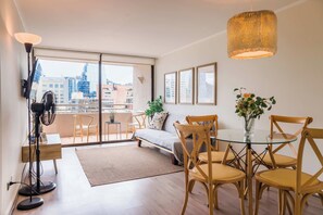 A modern living room with a balcony, a dining area with wooden chairs, a standing lamp, and framed artwork on the wall.