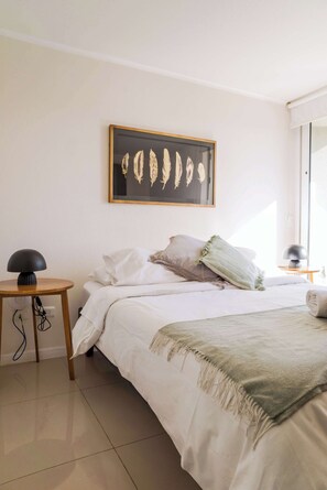 A bedroom with a bed, a bedside table with a lamp, a wall-mounted picture, and a window. - Your Santiago Retreat! Studio with Balcony & Wi-Fi (Santiago)