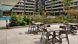 Un patio exterior moderno con varias mesas y sillas, rodeado de abundante follaje y una piscina.