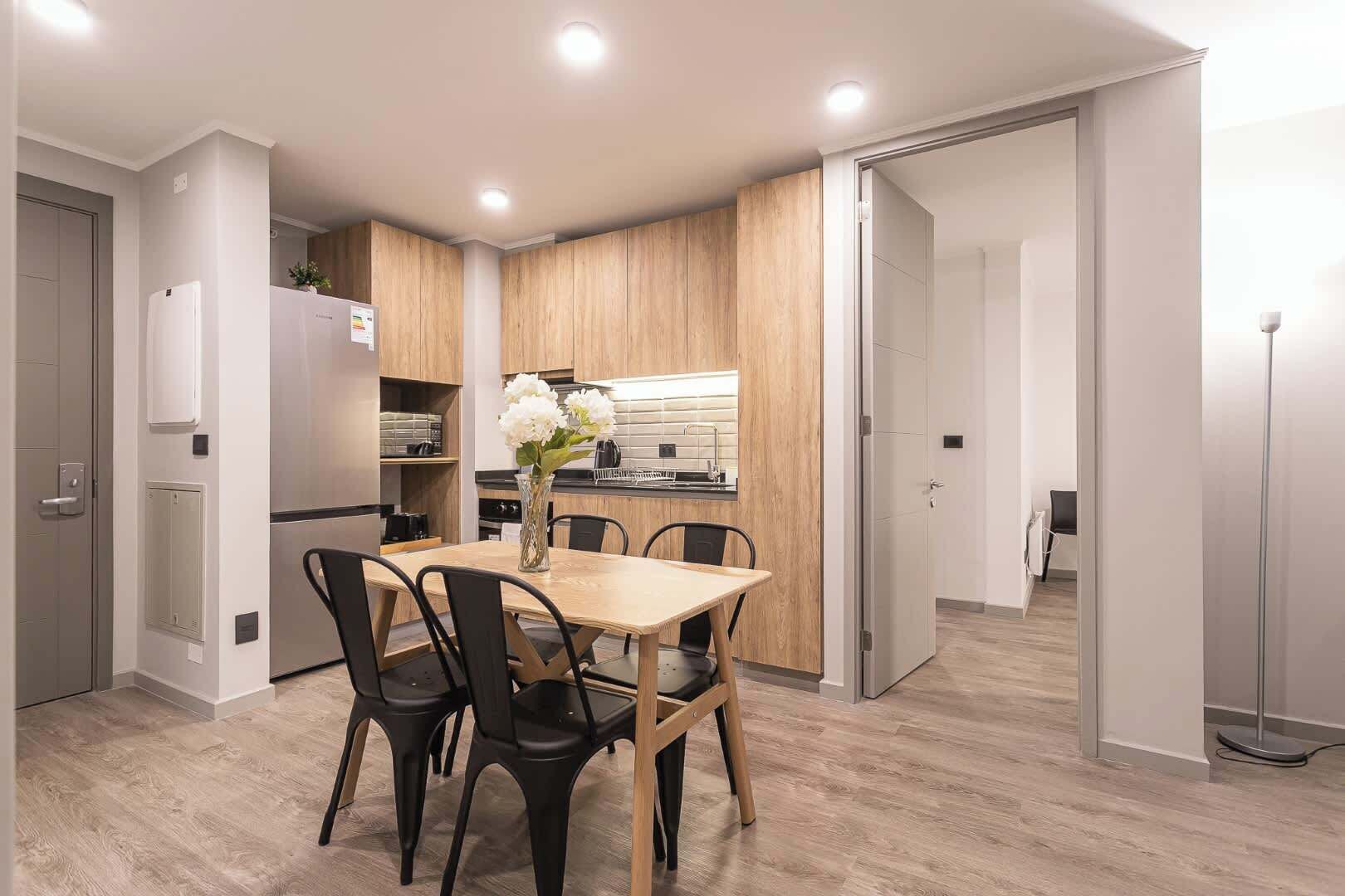 Een moderne keuken met houten kastjes, een eethoek met een tafel en stoelen, en een kleine koelkast.