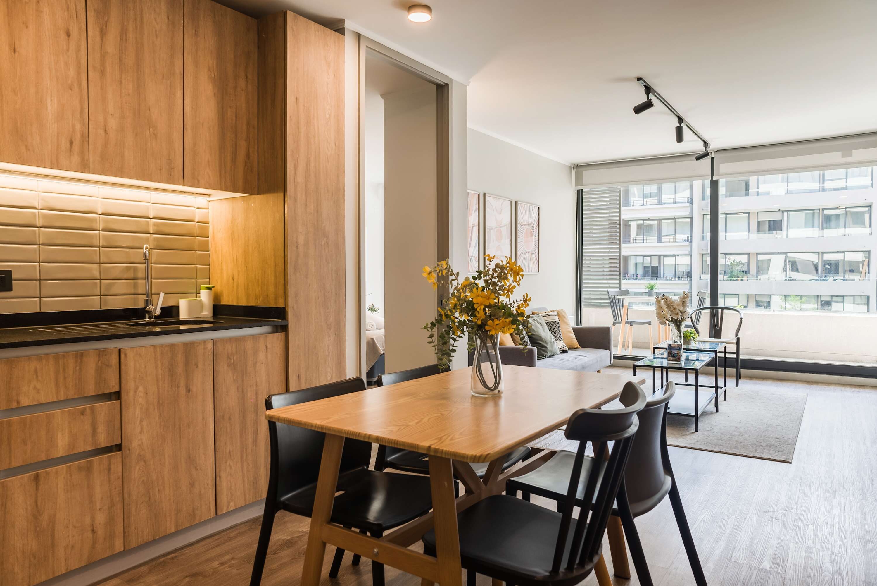 Una cocina moderna con armarios de madera y encimera negra, un comedor con mesa y sillas de madera, y ventanas amplias con vistas a la ciudad.