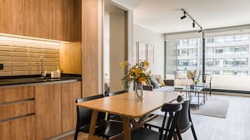 Una cocina moderna con armarios de madera y encimera negra, un comedor con mesa y sillas de madera, y ventanas amplias con vistas a la ciudad.