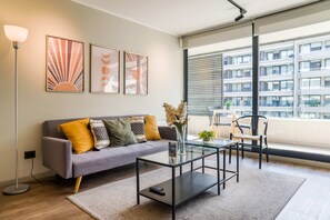 A modern living room with a grey sofa, a glass coffee table, and a floor lamp. There are three framed artworks on the wall and a large window with blinds. - Family Adventure Charming 2BR In Providencia For 5 (Providencia)