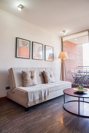 Un salon moderne avec un canapé, une table basse ronde et un lampadaire. Des œuvres d’art encadrées ornent le mur, et un mur de briques se trouve à l’arrière-plan.