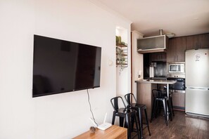 Une cuisine moderne équipée d’un téléviseur à écran plat fixé au mur, d’un four à micro-ondes et d’un réfrigérateur. On y trouve deux chaises noires et une petite table décorée d’un pommeau de pin.