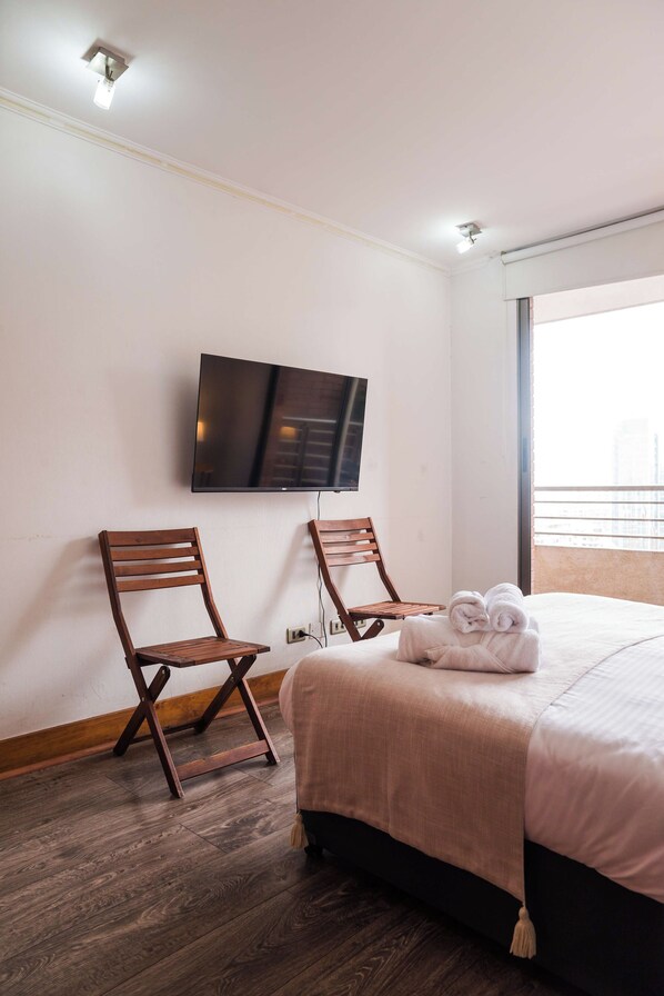 A hotel room with a bed, two wooden chairs, a flat-screen TV mounted on the wall, and a window with a view of buildings.