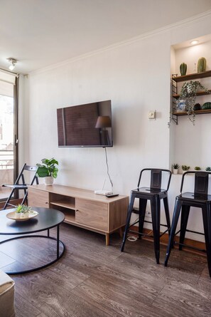 A modern living room with a flat-screen TV, a wooden entertainment unit, two black chairs, a round coffee table, and a potted plant. - Chic 2BR | Your Cozy Getaway Oasis in Las Condes (Las Condes)