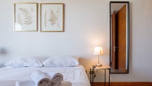 Ein ordentlich bezogenes Bett mit weißen Leinen, ein aufgerolltes Handtuch und zwei gerahmte Pflanzenbilder an der Wand.