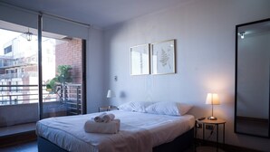 A hotel room with a bed, a nightstand, a lamp, a large window with a view of buildings, and a sliding door. - Cozy Apartment Perfect for Families and Long Stays (Las Condes)