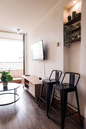 A modern living room with a flat-screen TV mounted on the wall, a wooden entertainment unit, two black chairs, a small round table with a potted plant, and a large window providing natural light.