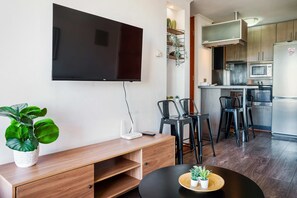 A modern living room with a flat-screen TV, wooden furniture, a dining area with chairs, and a kitchen with appliances. - Chic 2BR | Your Cozy Getaway Oasis in Las Condes (Las Condes)