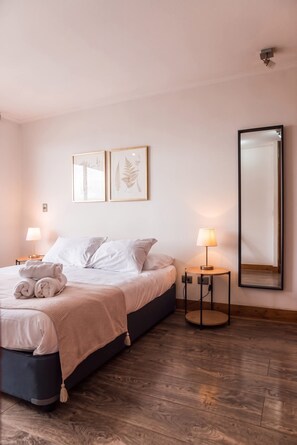 A bedroom with a bed, two bedside lamps, a round table with a lamp, a large mirror, and two framed botanical prints on the wall.