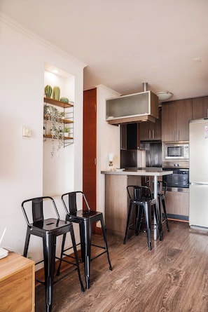 A modern kitchen with a built-in oven, microwave, and refrigerator. There are two black bar stools and a wooden dining table.