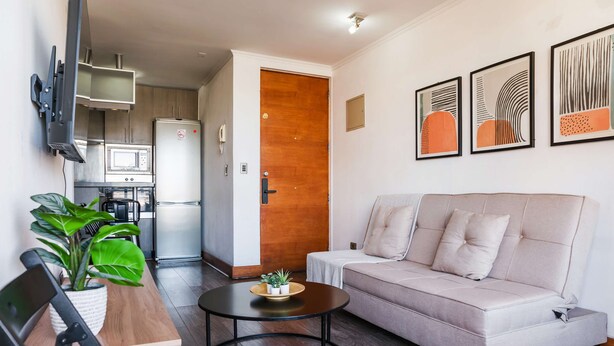 A modern living room with a sofa, a coffee table, a TV mounted on the wall, and a kitchen area visible through an open door. - Cozy Apartment Perfect for Families and Long Stays (Las Condes)