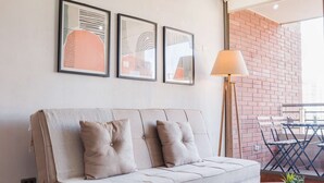 Ein modernes Wohnzimmer mit einer Couch, einem runden Couchtisch und einer Stehlampe. An der Wand befinden sich gerahmte Kunstwerke, und im Hintergrund ist eine Backsteinwand zu sehen.