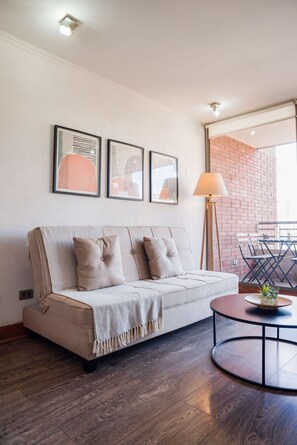 A modern living room with a sofa, a round coffee table, and a floor lamp. There are framed artworks on the wall and a brick wall in the background. - Chic 2BR | Your Cozy Getaway Oasis in Las Condes (Las Condes)
