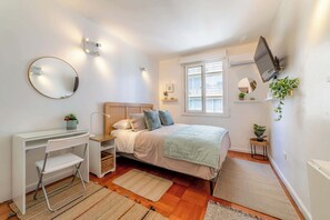 A bedroom with a bed, a desk, a chair, a TV, and a window.